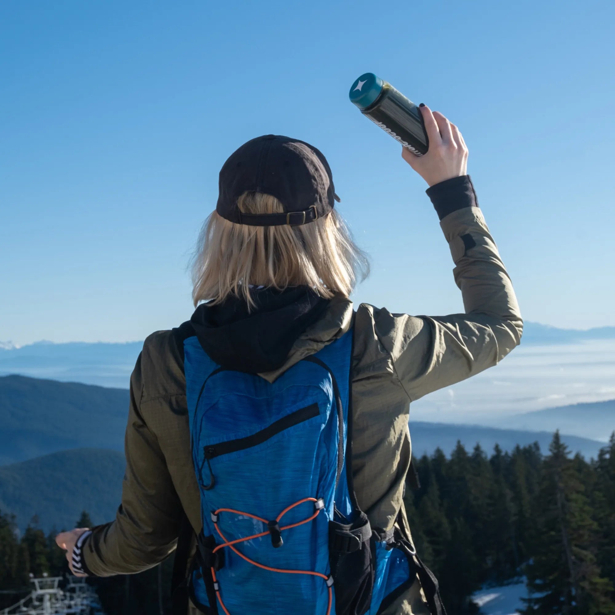 Person with a blue backpack taking a photo of a scenic mountain view and holding up a Magic Scoop Shaker Bottle.