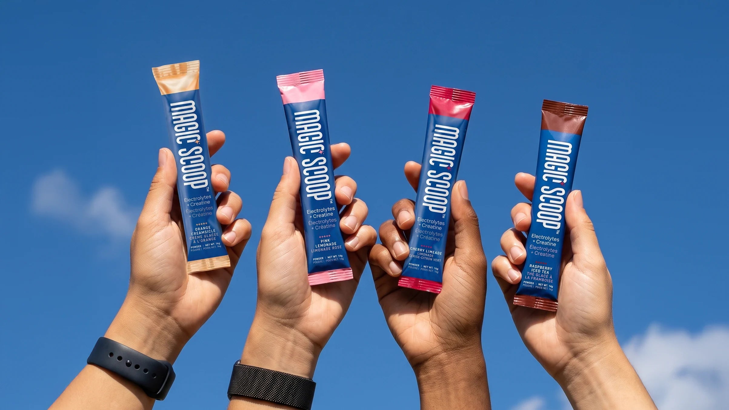 Four raised hands against a blue sky, each holding a Magic Scoop Electrolytes + Creatine stick pack: Orange Creamsicle, Pink Lemonade, Cherry Limeade and Raspberry Iced Tea.
